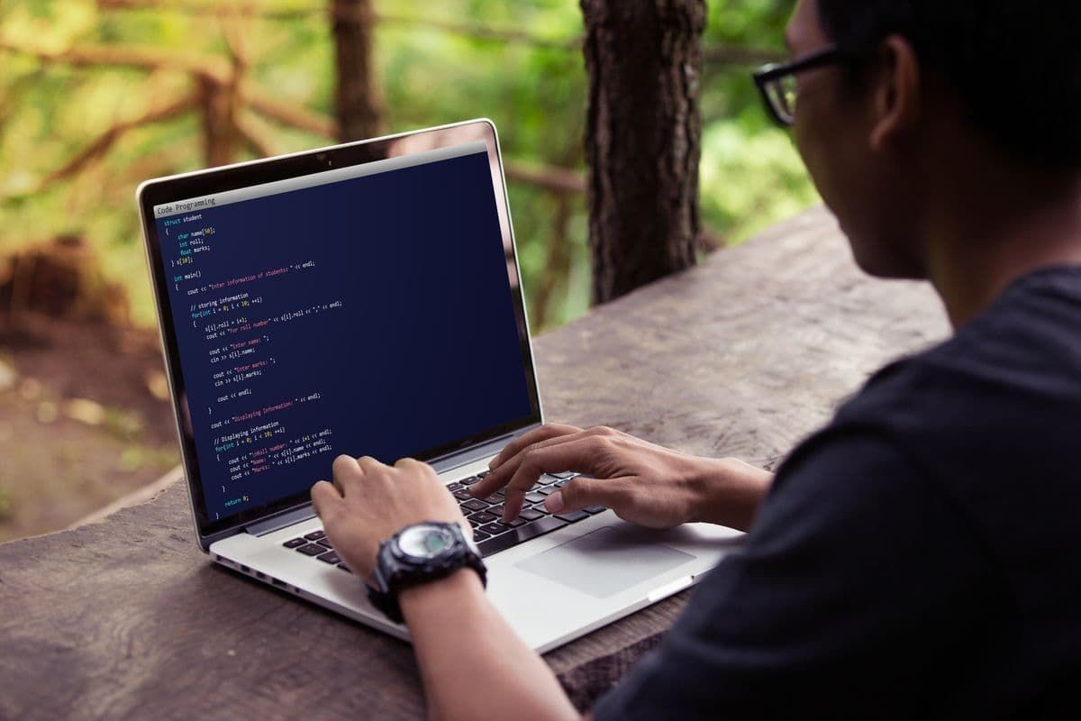 Person typing code on a laptop while sitting outdoors with a view of green trees in the background.