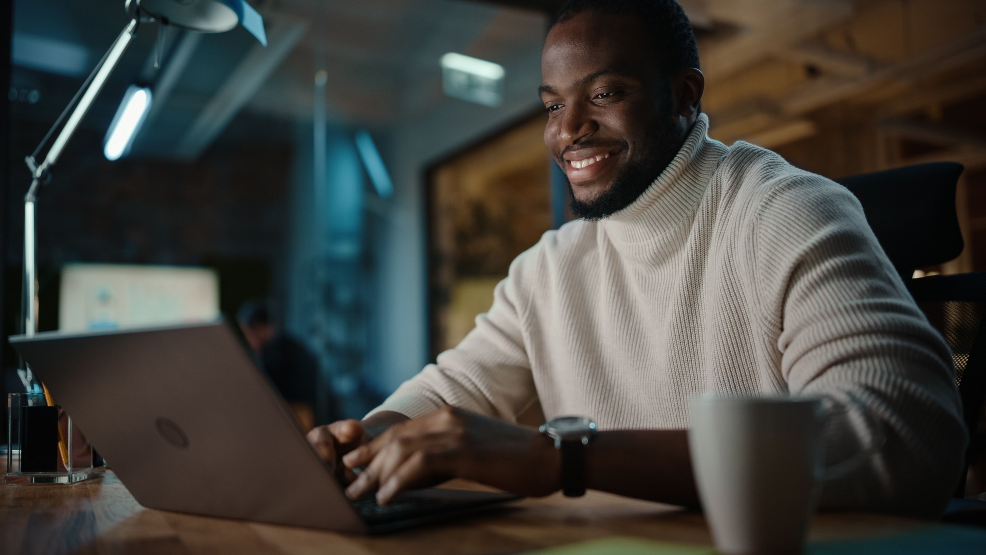 Bel homme afro-américain noir ayant une conversation en ligne sur un ordinateur portatif dans l’environnement créateur de bureau. Happy Male navigue sur les médias sociaux et répond à friends in Messenger.